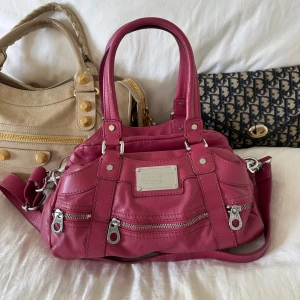 Rosa Marc by Marc Jacobs väska  - Stilren och tidlös Marc by Marc Jacobs Standard Supply väska i mjukt hallonrosa läder med silverfärgade detaljer. En eftertraktad modell som är både praktisk och elegant på samma gång. Perfekt vardagsväska som rymmer det nödvändigaste och passar lika bra till jobb som till fritid. Skriv för fler bilder privat och info🤍  Väskan har dubbla handtag och en avtagbar axelrem, så den kan bäras både i handen och crossbody. Framsidan pryds av två dragkedjefickor och den ikoniska logoplattan i metall. 