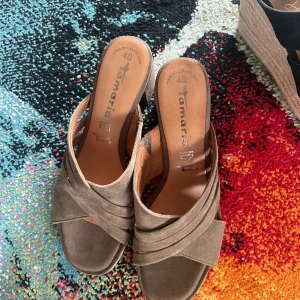 Bruna sandaler med kilklack från Tamaris - Snygga bruna sandaler från Tamaris med korslagda remmar i mocka och bekväm kilklack. Perfekta för sommaren och lätta att matcha med olika outfits. Skorna har öppen tå och en stilren design som passar till många tillfällen.