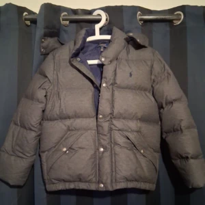 Grå dunjacka från Polo Ralph Lauren - Säljer en grå dunjacka från Polo Ralph Lauren med avtagbar huva och blått innerfoder. Jackan har tryckknappar, dragkedja och två snedställda fickor framtill. Klassisk Polo-logga på bröstet och Polo-detaljer längs dragkedjan. Perfekt för kalla dagar. Köpte den för några år sen och inte haft på mig den knappt ny pris 3000 kr .en liten skada men fixat så det inte syns,pris kan ändras vid snabb affär.