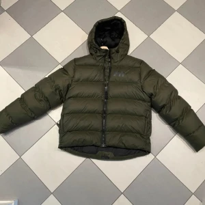 Dunjacka från Helly Hansen - Säljer en olivgrön pufferjacka från Helly Hansen med svart innerfoder och huva. Jackan har en tydlig HH-logga på bröstet, dragkedja framtill och breda, quiltade kanaler. Perfekt för kalla dagar och riktigt snygg streetstil.