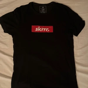 Svart t-shirt med röd skrrr-tryck - Svart t-shirt från FSBN i bomull med ett fett rött tryck på bröstet där det står 'skrrr.' i vitt. Klassisk rund hals och korta ärmar, perfekt för dig som vill sticka ut med streetstyle-vibe.