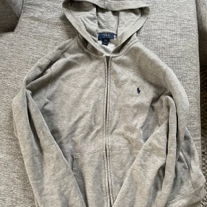Grå hoodie från Polo Ralph Lauren - Fin tröja men dragkedjan har gått sönder men går endå att använda. Finns några små defekter men som man inte ser. Finns en fläck i luvan men tror den går bort i tvätten har inte testat. Skriv för fler frågor