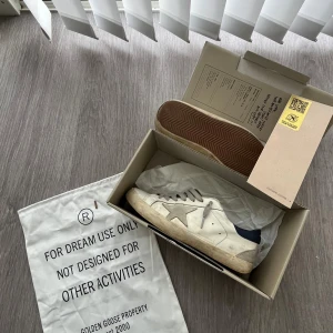 Golden Goose vit/blå - Säljer dessa feta golden goose i bra skick! De är vit med blå detalj vid hälen. Box plus distans medföljer och QR kod funkar Såkalrt. Enda defekten är en liten öppning på hälen på båda skorna. Men annars perfekta, plus super pris! Ny pris cirka 5000kr