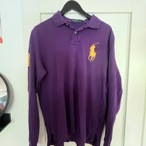 Lila långärmad pikétröja från Ralph Lauren - Säljer en lila långärmad pikétröja från Ralph Lauren med stor gul broderad logga på bröstet och gul detalj på ärmen. Klassisk krage och knäppning framtill. Perfekt för dig som vill sticka ut med en färgstark och stilren look. Storlek L, men funkar för mig som har M med lite ovetandes look.