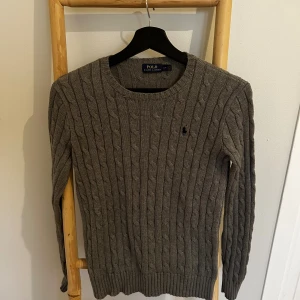 Grå kabelstickad tröja Polo Ralph Lauren - Kabelstickad grå tröja från Polo Ralph Lauren med rund halsringning och broderad logga på bröstet. Långärmad och i klassisk, tidlös design som är perfekt för lager-på-lager. Mjuk och skön känsla, passar till allt.