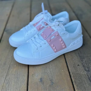 Valentino Rockstud sneakers vit/rosa - Snygga Valentino Garavani Rockstud sneakers i vitt skinn med ljusrosa detaljer och ikoniska nitar. Skorna har rund tå, platt sula och vita snören. Perfekta för dig som vill ha en lyxig och trendig look med en touch av rosa.