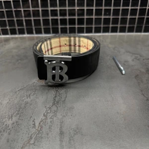 Hel svart bälte med silverbuckla  - Säljer ett snyggt bälte i svart skinn med klassiskt rutigt mönster på insidan och silverfärgat TB-spänne. Bältet har diskret logga och känns både stilrent och lyxigt. Perfekt accessoar för att lyfta din outfit.
