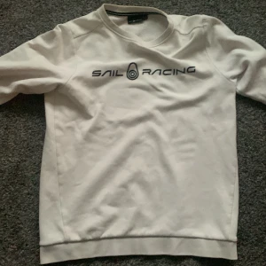 Vit Sail Racing tjocktröja  - Vit tjocktröja från Sail Racing med svart logga tryckt på bröstet. Tröjan har rund halsringning, långa ärmar och ribbade muddar vid ärmslut och nederkant. Perfekt för dig som gillar stilren och sportig look. Om man tittar riktig noga kan man se att den har några svarta pyttesmå prickar, men det är inget man märker av, därav det låga priset