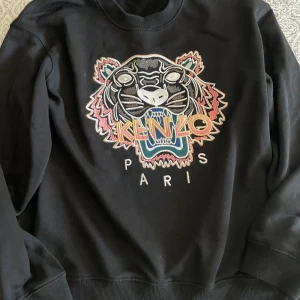 Svart Kenzo sweatshirt med tiger - Svart sweatshirt från Kenzo med ikoniskt broderat tigerhuvud och färgglad logga på bröstet. Tröjan har rund halsringning, långa ärmar och ribbade muddar. Perfekt statement-plagg för dig som gillar streetstyle och vill sticka ut.