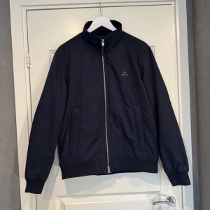 Marinblå vindjacka från GANT, M - Snygg marinblå vindjacka från GANT i klassisk modell med hög krage och silverfärgad dragkedja. Jackan har två fickor framtill, och två fickor på insidan, ribbade muddar och diskret logga på bröstet. Perfekt för vår och höst när du vill ha en stilren look.