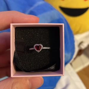 Pandora silverring med hjärta - Superfin silverring från Pandora med ett rött hjärta i mitten, omgiven av små gnistrande stenar. Ringen har en elegant och romantisk design som verkligen sticker ut. Perfekt för dig som älskar detaljer och vill ha något unikt på fingret.