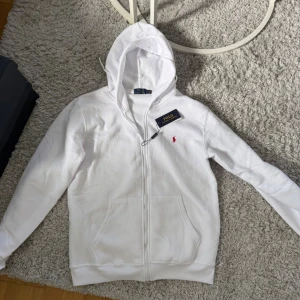 Vit hoodie från Polo Ralph Lauren - Vit hoodie från Polo Ralph Lauren med dragkedja och huva. Klassisk design med röd broderad logga på bröstet och två fickor framtill. Tillverkad i mjuk bomull, perfekt för chill dagar eller när du vill ha en clean look.