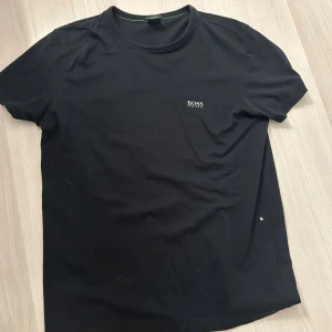 Svart t-shirt från Hugo Boss - Svart t-shirt från Hugo Boss med diskret logga på bröstet. Klassisk rund halsringning och korta ärmar. Tillverkad i mjuk bomull för en skön känsla. Perfekt basic-plagg med stilren design och snygga gröna detaljer på insidan av kragen. Och boss märke på ärmen.