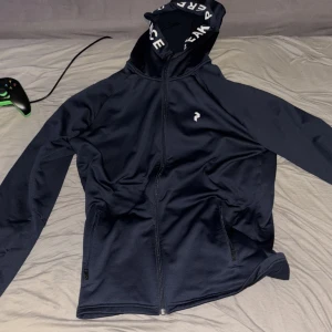 Marineblå hoodie från Peak Performance - Säljer en marineblå hoodie från Peak Performance med dragkedja och huva. Snygg logga på bröstet och text på insidan av huvan. Tröjan har långa ärmar och två fickor med dragkedja. Perfekt för dig som gillar stilren och sportig look.
