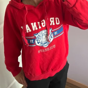 Röd hoodie med tryck och huva - Riktigt snygg röd hoodie med vit och blå text och en tiger på bröstet. Tröjan har huva med snörning och ribbade muddar. Perfekt för dig som gillar sportig och avslappnad stil. Materialet känns mjukt och skönt, troligen bomull eller bomullsblandning.