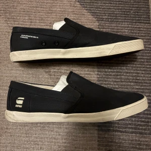 Svarta loafers från G-Star RAW - Svarta slip-on sneakers från G-Star RAW med vit sula och diskret logga på hälen. Skorna är i canvasmaterial och har en klassisk, låg profil med rund tå. Insidan har ett coolt mönster i blått och vitt. Perfekta för en avslappnad streetstil. Nypris ca 1200