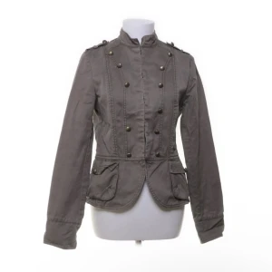 Grå militärinspirerad kavajjacka - Snygg grå kavajjacka med militärvibbar, dekorerad med dubbla rader metallknappar framtill och axelklaffar. Jackan har figurnära passform, två fickor med lock och hög krage. Perfekt för dig som vill sticka ut med en cool och stilren look.