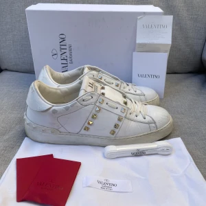 Valentino skor - Säljer ett par vita Valentino Garavani Rockstud sneakers i skinn med ikoniska guldiga nitar och snörning. Klassisk rund tå och platt sula, detaljerade med märkets logga på plösen. Snygga och lyxiga sneakers som sticker ut med sina metalldetaljer. Dustbag och kvitto medföljer