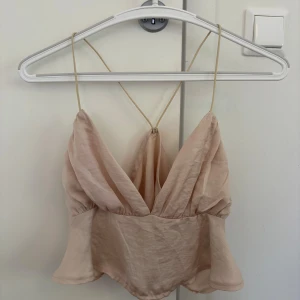 Beige linnetopp med tunna axelband - Säljer en ljusbeige linnetopp med djup v-ringning och tunna axelband som korsas i ryggen. Toppen har en lätt utsvängd nederdel och är tillverkad i ett tunt, luftigt material. Perfekt för varma dagar och en stilren look.