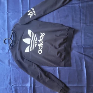 Svart Adidas sweatshirt med tryck - Svart sweatshirt från Adidas med stort vitt logotryck på bröstet och mindre logga på ena ärmen. Tröjan har rund halsringning och långa ärmar med muddar. Perfekt för en sportig och avslappnad stil.