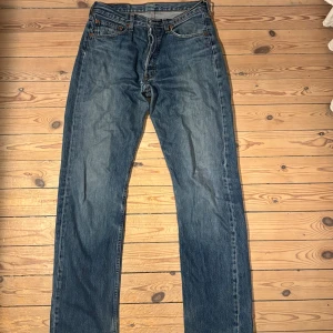 Levi's 501 blå jeans straight fit - Klassiska Levi's 501 jeans i blå tvätt med raka ben och fem fickor. Jeansen har knappgylf, orange sömmar och den ikoniska läderpatchen bak i midjan. Materialet är slitstarkt denim i bomull och passformen är normal med straight fit.