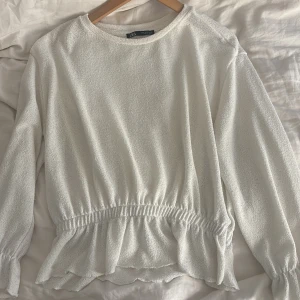 Vit blus med volang från Zara - Superfin vit blus från Zara med rund halsringning och långa ärmar. Blusen har en markerad midja med volangkant nedtill som ger en söt och trendig look. Materialet är lätt strukturerat och känns mjukt mot huden. Perfekt att styla med jeans eller kjol.
