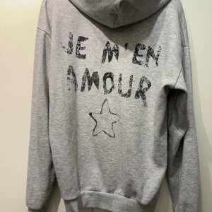 Grå hoodie från Gina Tricot  - Grå hoodie från Gina Tricot med svart texttryck 'Je m'en amour' och en stjärna både fram och bak. Använd typ 3 gånger i storlek 158-164 men käns större