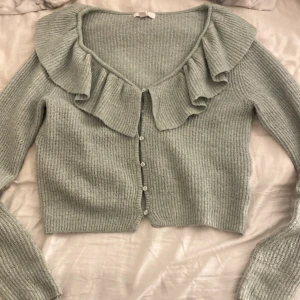 Ljusgrön stickad kofta med volang - Superfin ljusgrön stickad kofta med volangdetalj längs halsringningen och knappar framtill. Modellen är croppad och har långa ärmar. Perfekt för dig som gillar söta detaljer och vill ha en mysig men stylish look.