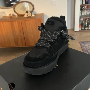 Svarta lowmells - Säljer ett par svarta boots med grov platt sula och breda snören i svart och grått mönster. Skon har detaljer i mocka och textil samt vadderad krage för extra komfort. Perfekt för dig som vill ha en chunky och stilren look.