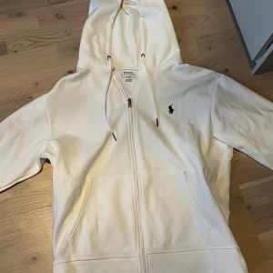 Vit hoodie från Polo Ralph Lauren - Vit hoodie från Polo Ralph Lauren med klassisk svart broderad logga på bröstet. Tröjan har huva med snörning, dragkedja framtill och två stora fickor. Tillverkad i mjuk bomull, perfekt för chill dagar. Snygg och enkel design som alltid funkar.