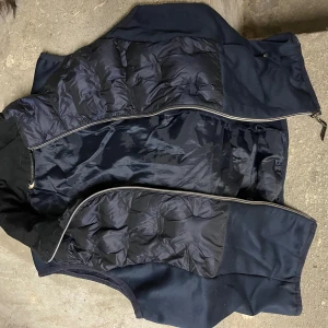 Mörkblå dunväst från JH Collection - Snygg mörkblå dunväst från JH Collection med quiltade partier och dragkedja framtill. Västen har en stilren look med blandning av glansigt och matt tyg, perfekt för lager-på-lager. Passar dig som gillar klassisk och sportig stil.