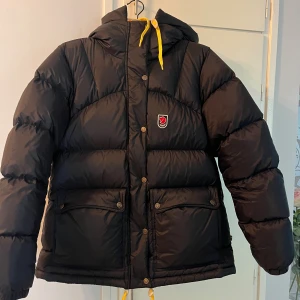 Mörkblå Fjällräven Expedition dunjacka - Mörkblå  Fjällräven Expedition Series dunjacka med huva och gula snören. Jackan har två stora fickor med knappar, broderad logga på bröstet och dragkedja med knappslå. Perfekt för kalla vinterdagar och har en riktigt puffig look.