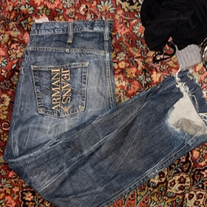 Armani Jeans med slitningar - Säljer ett par blå jeans från Armani Jeans med stor logga broderad på bakfickan. Jeansen har slitningar och hål vid knät för en cool, avslappnad look. Klassisk femficksmodell och normal passform.