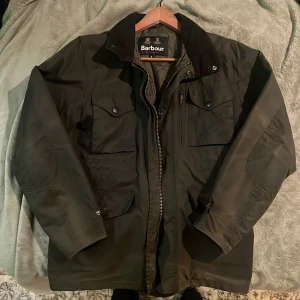 Mörkgrön Barbour Sapper jacket  - En klassisk mörkgrön vaxad jacka från Barbour, modell Sapper, i väldigt bra skick! Jackan har flera praktiska fickor med knappar, quiltat foder och dragkedja framtill. Perfekt för dig som gillar tidlös stil och funktion. Funkar bra till vintern och hösten. Kragen är i manchester och går att knäppa hela vägen upp, det finns en luva i kragen bakom en dold dragkedja som man kan väckla ut . Priset går att diskutera