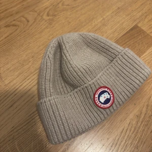 Beige ribbstickad mössa från Canada Goose - Ribbstickad beige mössa från Canada Goose med klassisk rund logotyp framtill. Tillverkad i 100% ull och har en bred uppvikt kant för extra värme. Perfekt för kalla dagar och snygg till alla vinteroutfits.