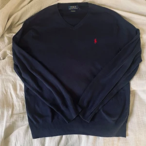 Mörkblå tröja från Ralph Lauren - Stilren tröja från Ralph Lauren i mörkblått med v-ringning och den klassiska röda loggan på bröstet. En tidlös modell som alltid funkar – både till vardags och mer dressat.  📏 Storlek: L ✨ Skick: Bra skick, välvårdad och utan defekter. 🧵 Material: Bomullsmix – mjuk, lätt och bekväm.        ✅ Pris kan diskuteras