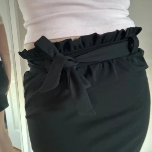 Svart kortkjol med volang och knyt - Säljer en svart kortkjol med snygg volangkant upptill och ett brett knytband i midjan. Kjolen har en rak passform och är tillverkad i mjukt tyg som sitter skönt. Perfekt för dig som gillar stilrena plagg med en twist. Plagget är från Gina tricot och i mycket bra skick.