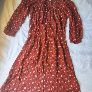Rostfärgad blommig klänning med knyt stl 32/34 - Superfin rostfärgad klänning med små vita och gula blommor, långa ärmar och smockdetaljer vid axlar och midja. Klänningen har knytband vid halsen och är tillverkad i ett mjukt, lätt tyg som ger en avslappnad vibe.