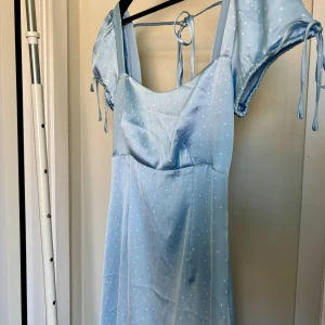 Ljusblå prickig klänning  - Superfin ljusblå klänning med vita prickar och puffärmar med knytband. Klänningen har en söt ringning, knyt i ryggen och är tillverkad i ett glansigt, mjukt material som känns lyxigt. Perfekt för dig som gillar romantisk och trendig stil. Från NAKD, aldrig använd därav prislapp kvar. Från kollektionen Ida Carlsson x NAKD.