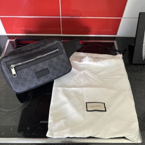 Svart magväska  med monogram - Snygg svart axelväska från med klassiskt monogram i grått. Väskan har silverfärgad dragkedja, justerbart band i rött och blått samt detaljer i skinn. Perfekt storlek för det viktigaste och levereras med original dustbag.