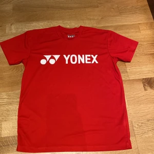 Yonex - Röd  t-shirt med tryck - Badminton  - Röd t-shirt från Yonex med vitt logotryck på bröstet. Klassisk rund hals och korta ärmar. Perfekt för dig som gillar sportig stil och vill sticka ut med en färgstark look.
