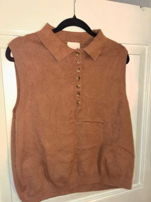 Brun stickad väst med krage H&M - Säljer en brun stickad väst från H&M med klassisk krage och snygga knappar framtill. Västen är ärmlös och har en rak passform som är enkel att styla över en skjorta eller t-shirt. Perfekt för lager-på-lager-look under höst och vinter.