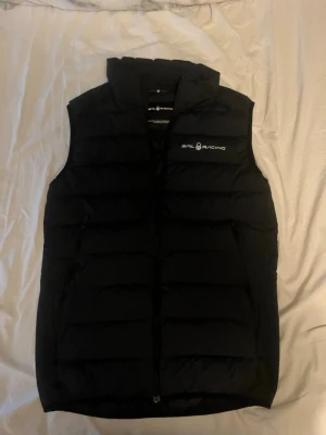 Svart dunväst från Sail Racing - Säljer en svart dunväst från Sail Racing med hög krage och dragkedja framtill. Västen har två sidofickor med dragkedja och diskret logga på bröstet. Fylld med Allied Feather & Down, 800 fill power. Perfekt för lager-på-lager under kyliga dagar.