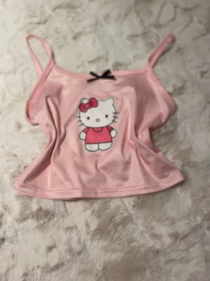 Hello Kitty rosa linnetopp XS - Supersöt rosa linnetopp med Hello Kitty-tryck framtill och en liten svart rosett vid halsringningen. Toppen har smala axelband och croppad passform, perfekt för en gullig och lekfull stil. Materialet känns mjukt och stretchigt.