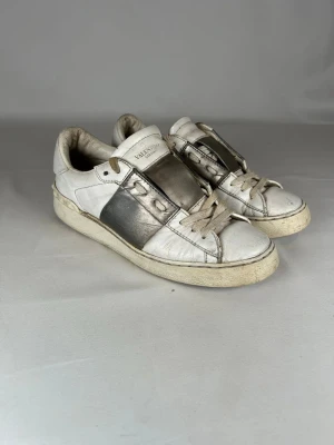 Valentino Garavani vita gråa - (Nypris 6200kr)Snygga vita sneakers från Valentino Garavani med metallic-silver detalj på sidan och klassisk snörning. Skorna är tillverkade i skinn och har en platt sula samt rund tå. Perfekta för dig som gillar exklusiva och trendiga märken. (Size 37,5 men passar även 38)