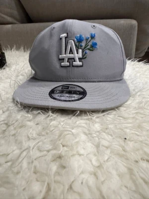 New Era Los Angeles Dodgers Flower Icon Grey 9FIFTY Snapback - Modell: New Era 9FIFTY 	•	Lag/logga: LA Dodgers 	•	Färg: Grå 	•	Detalj: Blå blommig brodyr bredvid LA-loggan 	•	Stängning: Snapback (justerbar)