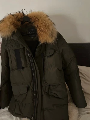 Hollies jacka  - Säljer en olivgrön parkas från Hollies i storlek XL. Jackan har en stor fluffig pälsimitation på huvan, flera praktiska fickor och dragkedja framtill. Perfekt för kalla dagar med sin rejäla look och snygga detaljer.