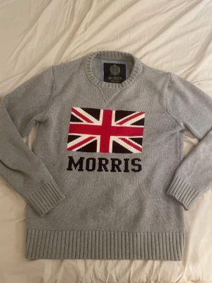 Grå stickad tröja Morris med flagga - Snygg grå stickad tröja från Morris med Union Jack-flagga och stor logga framtill. Tröjan har rund hals, ribbade muddar och är långärmad. Perfekt för dig som gillar klassisk brittisk stil och vill sticka ut lite extra.
