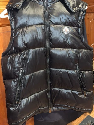 Svart dunväst från Moncler - Säljer en svart dunväst från Moncler med glansig finish och huva. Västen har två fickor med dragkedja och klassisk Moncler-logga på bröstet. Insidan har ett unikt serietidningsmärke. Perfekt för dig som vill ha en cool och varm look.