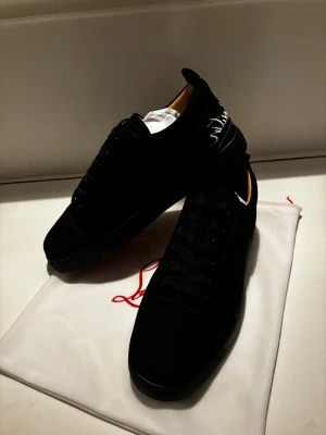Svarta sneakers från Louboutin - Snygga svarta sneakers från Christian Louboutin i mocka med klassisk röd sula och vit broderad logga på hälen. Skorna har rund tå, svarta snören och diskreta detaljer som ger en lyxig vibe. Perfekta för dig som vill sticka ut med exklusiva skor. Med följer dock ingen låda
